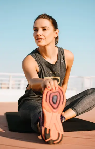 Mujer haciendo estiramientos sentada al aire libre, vestida con ropa deportiva y sonriendo.