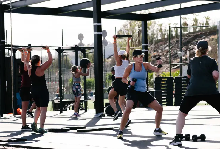 Grupo de personas haciendo ejercicio al aire libre con pesas y bandas elásticas en un gimnasio exterior.