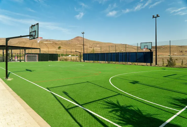 Cancha deportiva al aire libre con césped artificial, aros de baloncesto y porterías, rodeada de montañas.