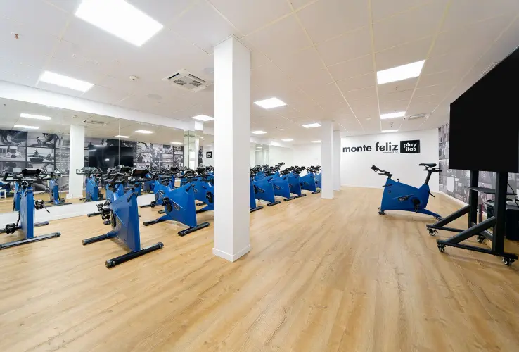 Sala de spinning con bicicletas estáticas azules, suelo de madera, espejos y un monitor grande al fondo.