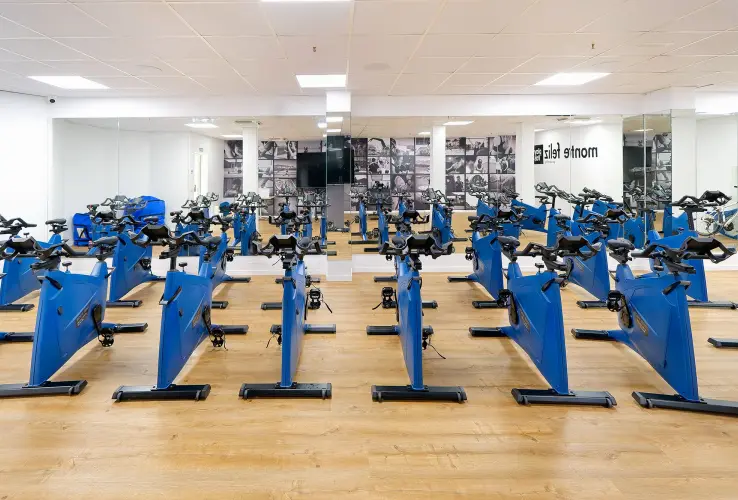 Sala de spinning con varias bicicletas estáticas azules, suelo de madera y paredes con espejos.
