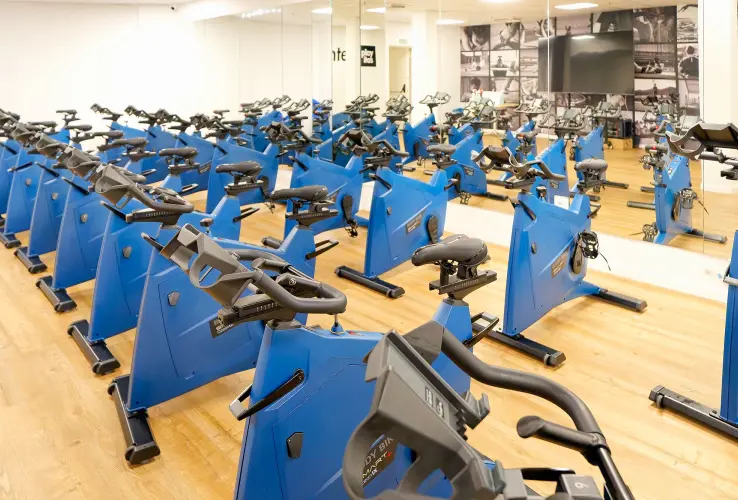 Sala de spinning con numerosas bicicletas estáticas azules en un gimnasio, sobre suelo de madera.