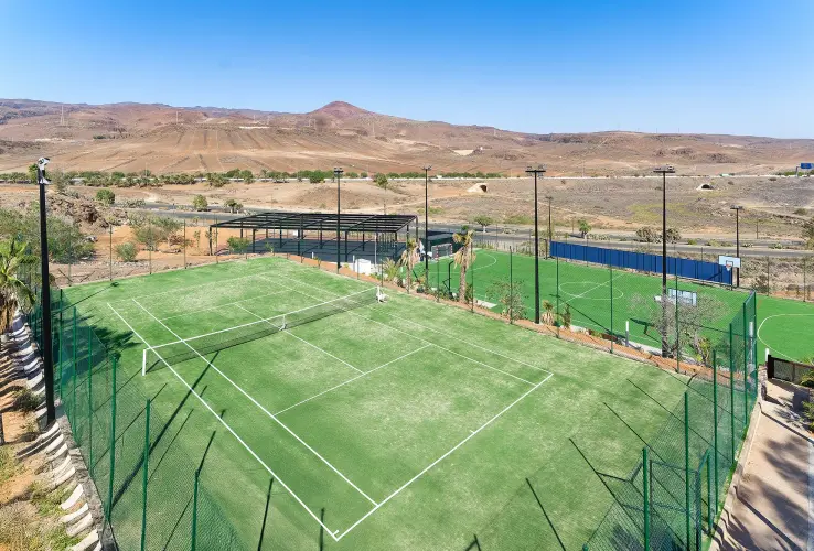 Cancha de tenis al aire libre rodeada de una cerca, con paisaje árido y colinas al fondo bajo un cielo despejado.