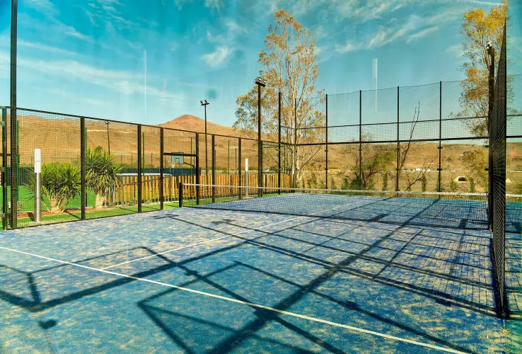 Pista de pádel al aire libre con suelo azul y vallas metálicas, rodeada de paisaje montañoso y árboles.