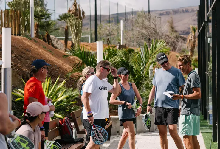 Grupo de personas con palas de pádel conversando al aire libre, rodeados de vegetación y una cancha al fondo.
