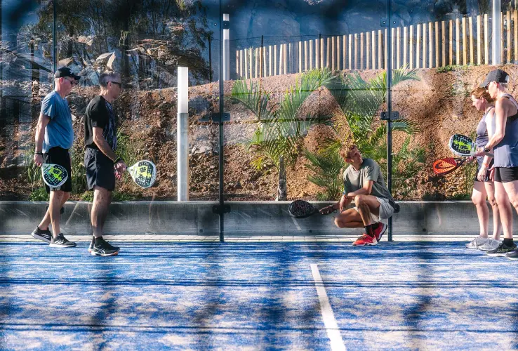 Personas jugando pádel en una cancha azul, rodeados de una pared de vidrio con vegetación al fondo.