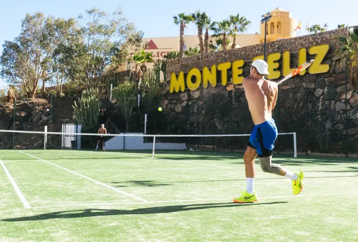 Jugador de tenis en acción en una cancha exterior bajo el sol, con "MONTE FELIZ" en el fondo.