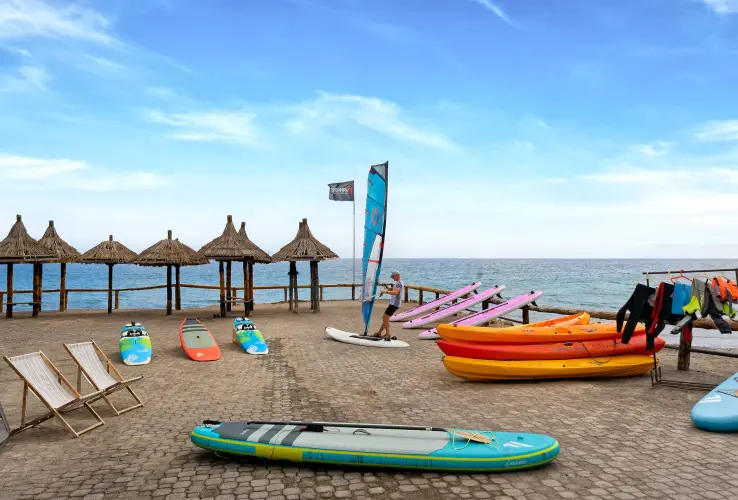 Playa con sombrillas de paja, tablas de surf, y kayaks coloridos en la arena junto a un mar azul bajo un cielo despejado.