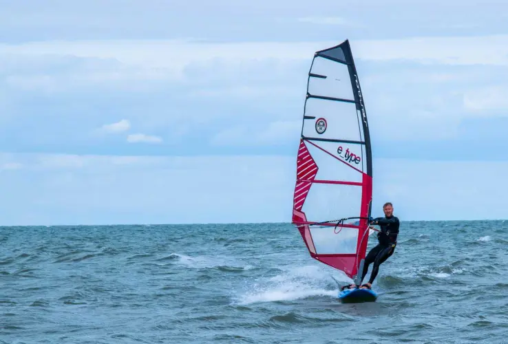 Una persona practica windsurf en el mar, con una vela roja y blanca y el cielo nublado al fondo.