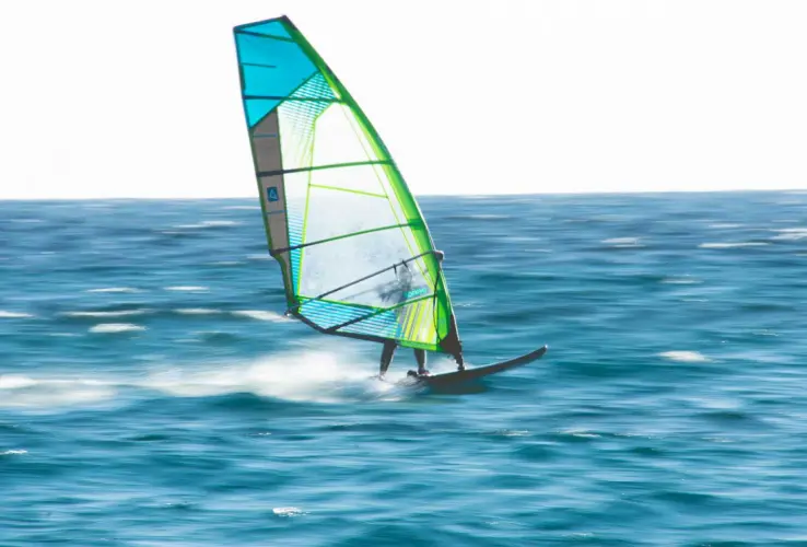 Windsurfista sobre azul intenso del océano, con vela verde y azul, moviéndose rápidamente sobre el agua.