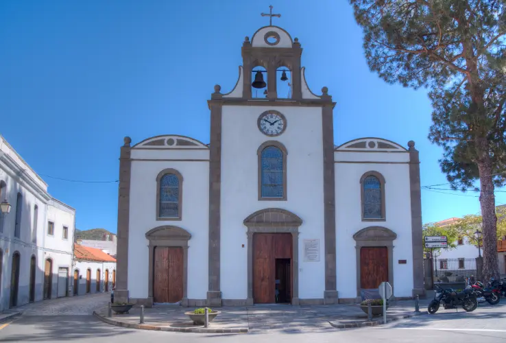Fachada de una iglesia con campanario, relojes y puertas marrones, rodeada de edificios y un árbol grande a la derecha.