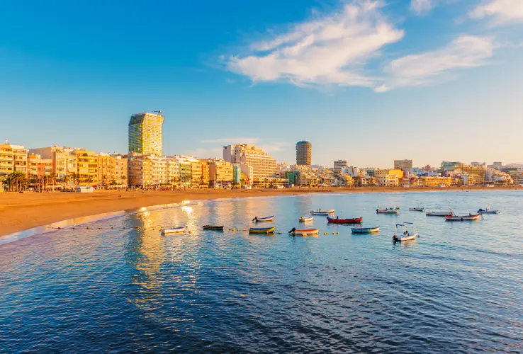 Playa con barcos y edificios modernos al atardecer bajo un cielo ligeramente nublado.