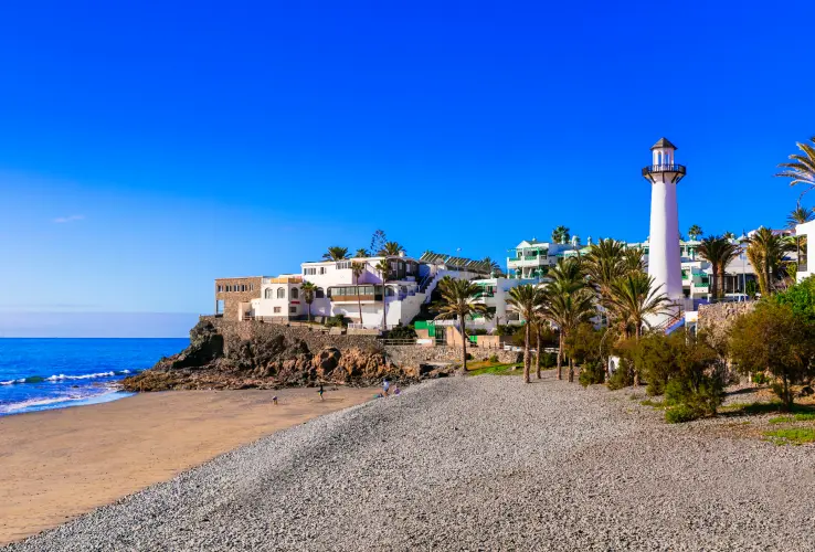 Playa con faro y casas blancas al fondo, rodeada de palmeras, bajo un cielo azul despejado.