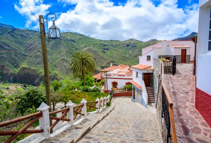 Calle empedrada en un pueblo con casas blancas y techos rojos, rodeada de montañas y vegetación, con cielo azul y nubes.