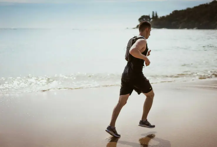 Persona corriendo en la playa junto al mar, con un chaleco y zapatillas deportivas bajo un cielo despejado.