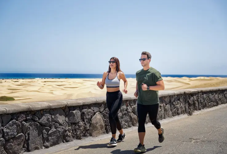 Dos personas corren juntas por una carretera junto a dunas de arena y el mar al fondo, bajo un cielo despejado.