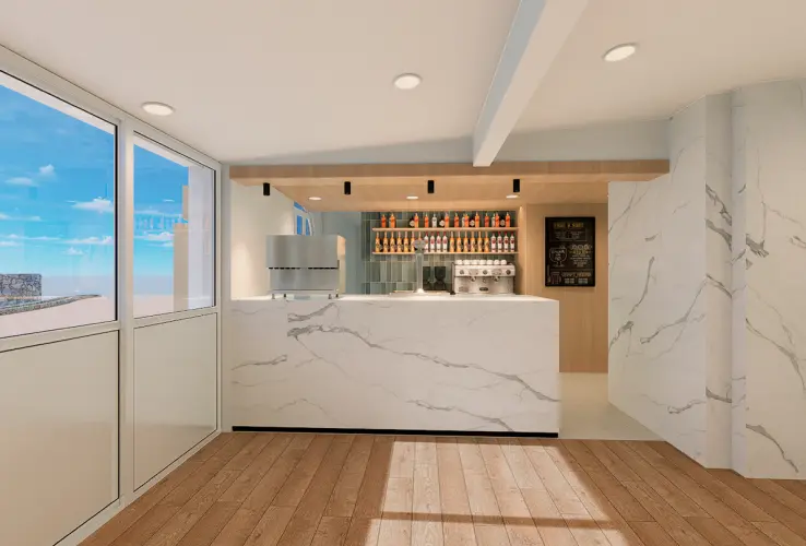 Cafetería moderna con mostrador de mármol blanco, estantes de bebidas y piso de madera, junto a grandes ventanas.