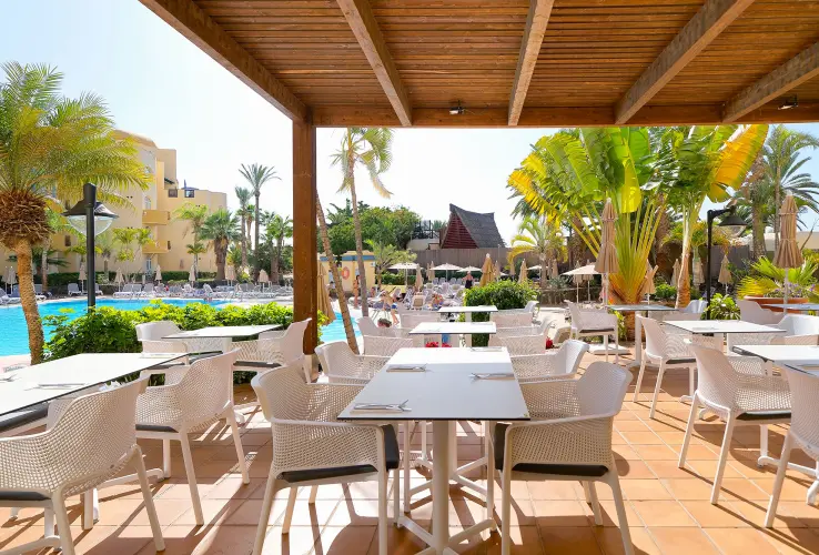 Área de comedor al aire libre con mesas y sillas blancas junto a una piscina rodeada de palmeras y tumbonas.