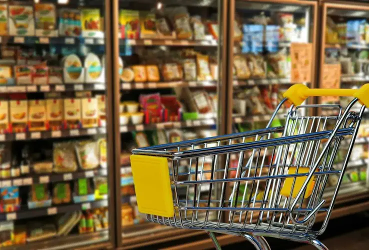 Carrito de compras vacío frente a estantes llenos de productos en un supermercado.