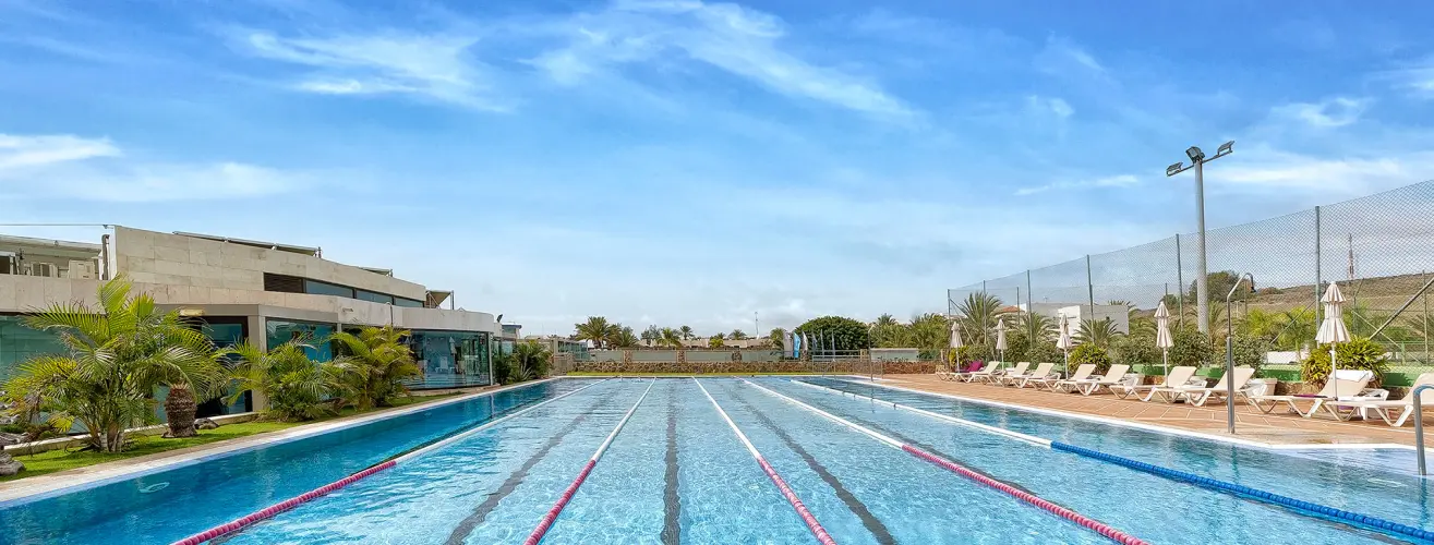 Piscina olímpica al aire libre, con carriles marcados, rodeada de palmeras y tumbonas bajo un cielo despejado.
