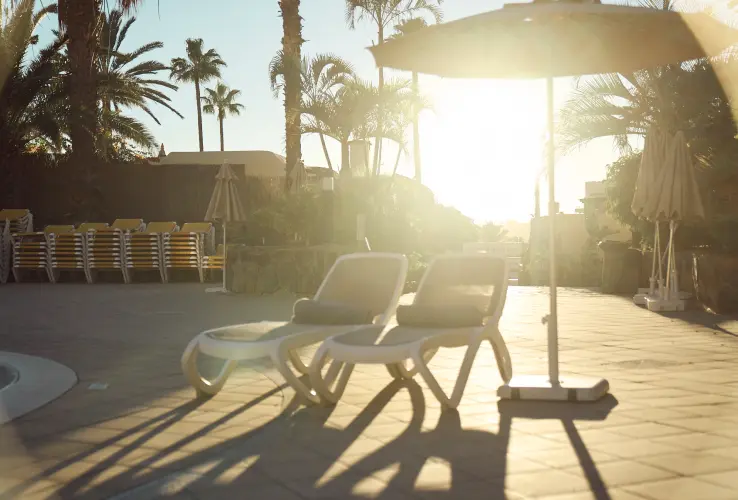 Sillas reclinables bajo una sombrilla junto a la piscina al atardecer, rodeadas de palmeras.