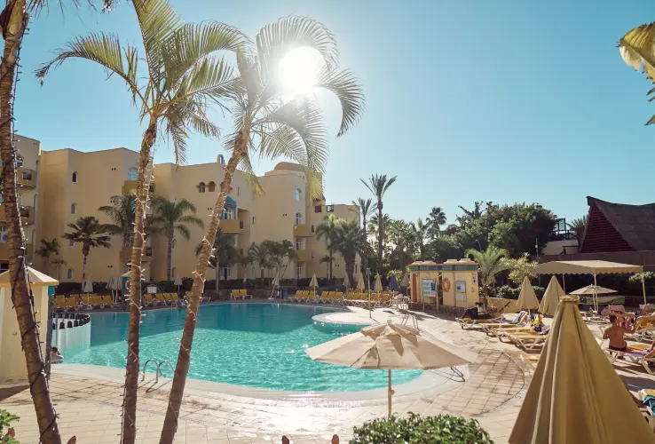 Piscina rodeada de palmeras y tumbonas bajo el sol brillante, con un hotel amarillo al fondo.