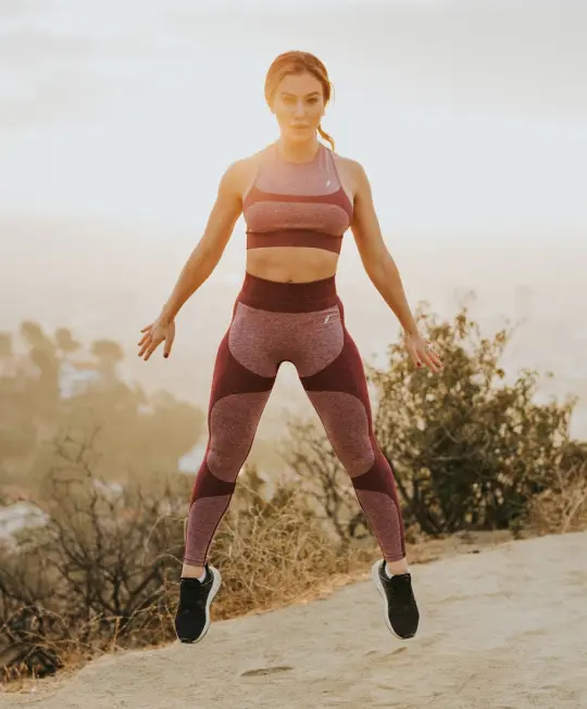 Mujer en ropa deportiva morada haciendo ejercicio al aire libre al amanecer, rodeada de vegetación y paisaje difuso.