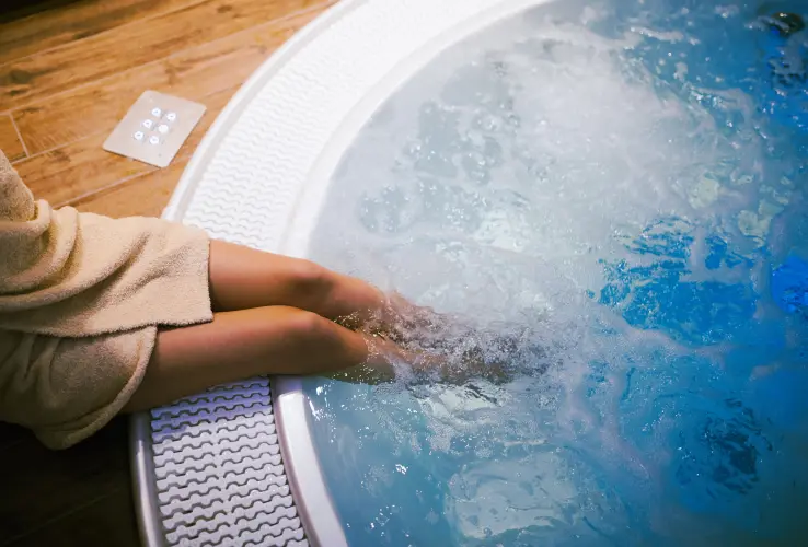 Persona con bata sentada al borde de un jacuzzi con las piernas dentro del agua, en un ambiente de spa.
