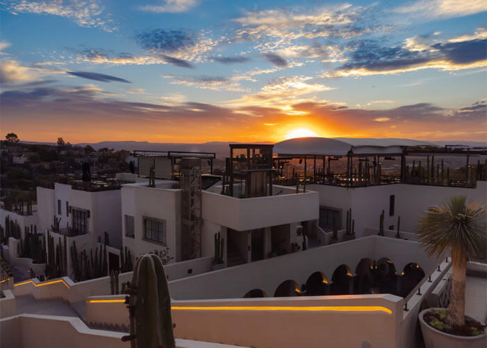 Edificios blancos rodeados de cactus al atardecer, con un cielo azul y anaranjado en el horizonte.