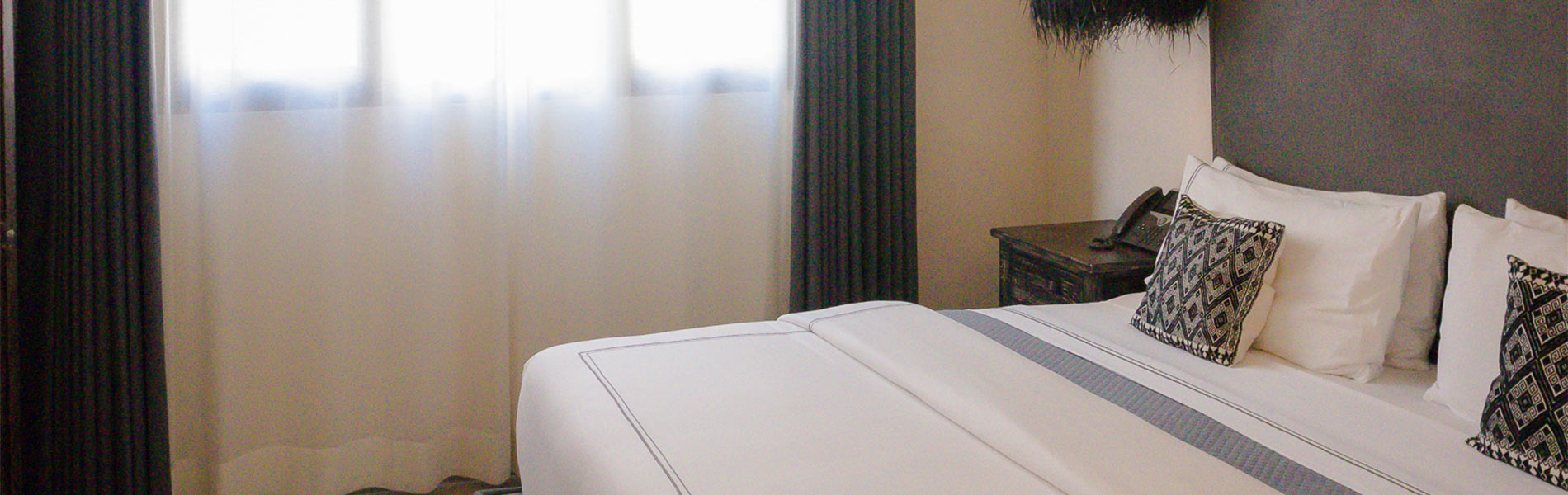Cama con sábanas blancas, cojines decorativos y cortinas oscuras en una habitación iluminada.