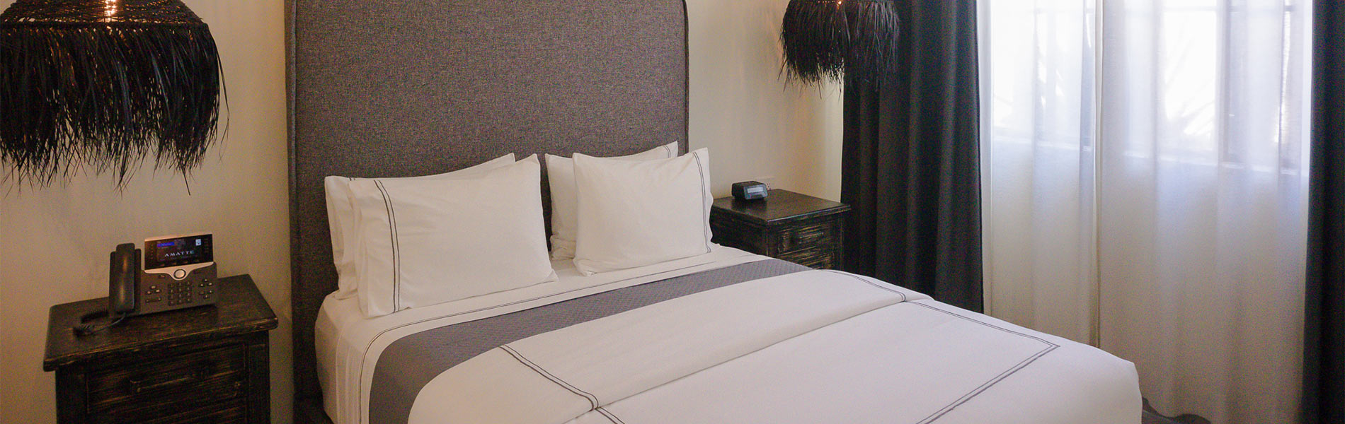 Habitación de hotel con cama doble, sábanas blancas, lámparas colgantes y cortinas oscuras junto a una ventana.