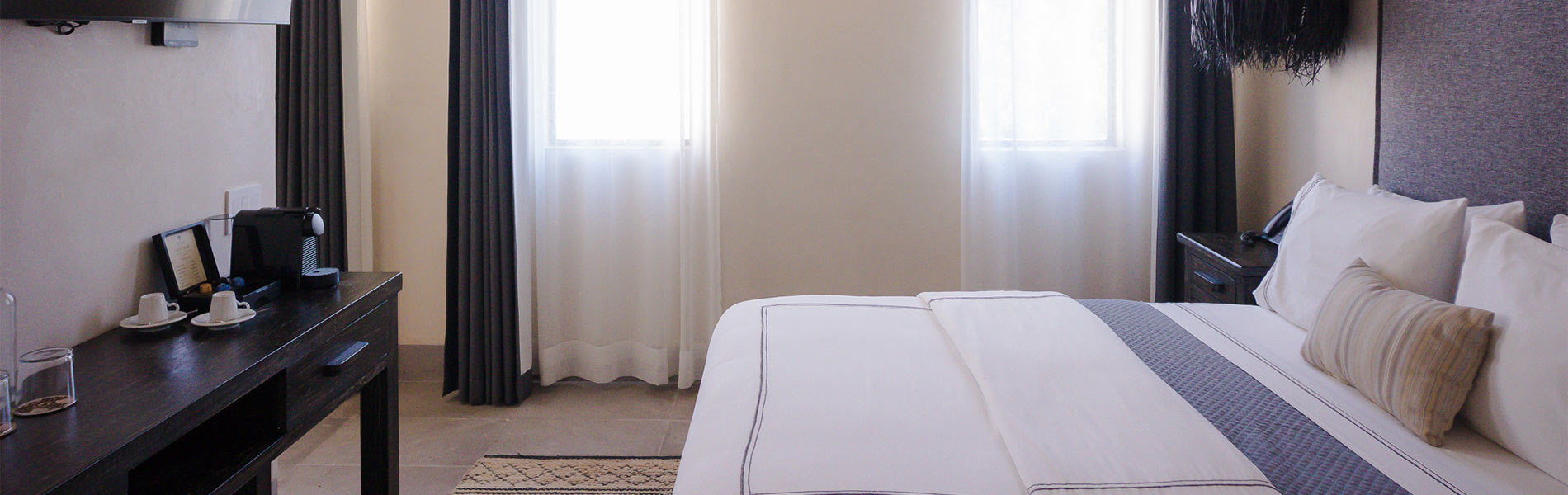 Habitación de hotel moderna con cama doble, cortinas oscuras y mesa con cafetera y tazas.