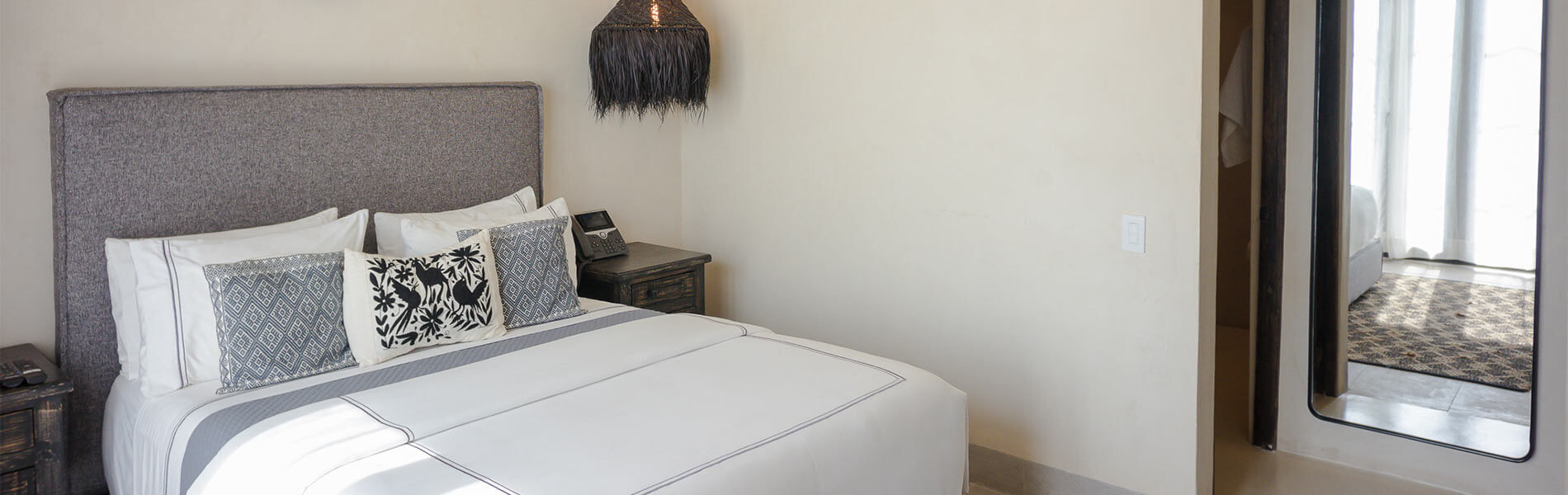 Habitación con cama doble, cojines decorativos, lámpara colgante, espejo alto y luz natural entrando por la ventana.