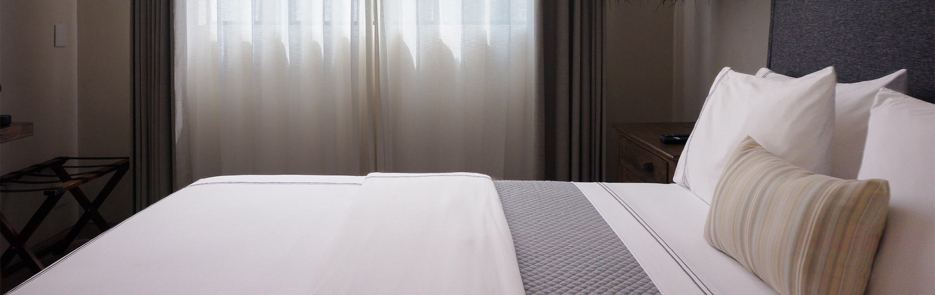 Habitación con cama de sábanas blancas, almohadas y ventana con cortinas translúcidas.