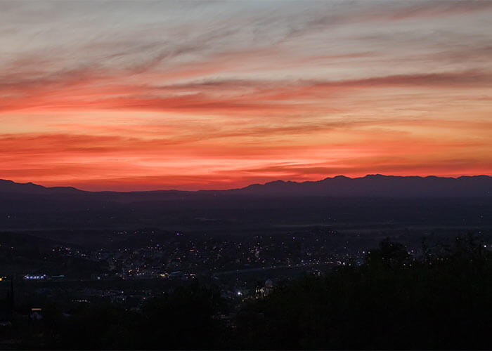 Atardecer rojo sobre montañas con un cielo parcialmente nublado y una ciudad iluminada abajo.