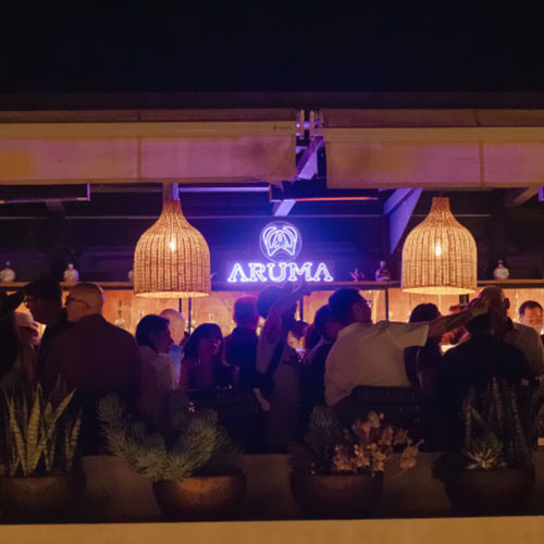 Personas socializando en un bar nocturno iluminado con luces colgantes y un letrero que dice "Aruma".