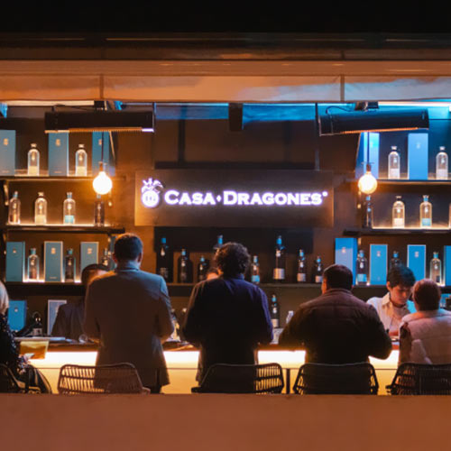 Personas en un bar iluminado con estantes llenos de botellas detrás del mostrador y un letrero de "Casa Dragones".