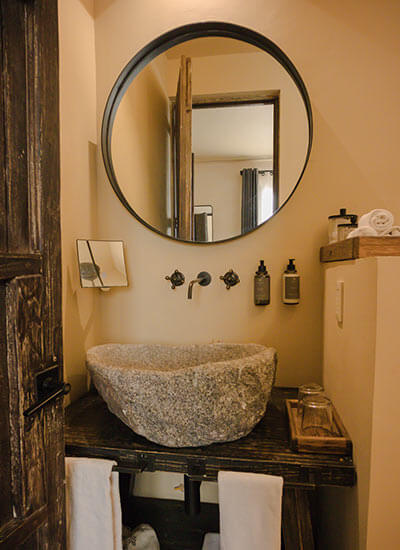Baño rústico con lavabo de piedra, espejo redondo, grifería de pared y accesorios de baño sobre una repisa.