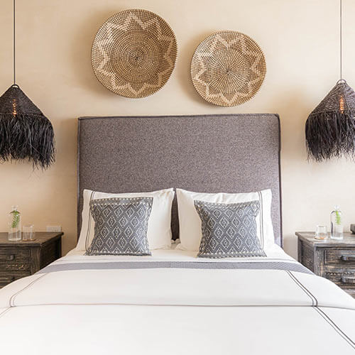 Cama con cabecero gris y cojines decorativos, dos cestas en la pared y lámparas colgantes negras a los lados.