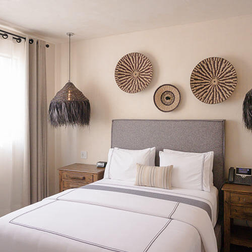 Dormitorio con cama doble, cabecera gris, cojines, lámpara colgante, mesas de noche y decoración de pared con cestas circulares.
