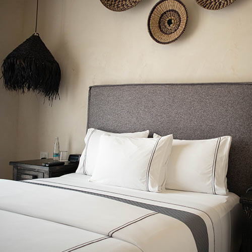 Habitación con cama de cabecera gris, cojines blancos, mesa de noche con lámpara negra y decoración de pared tejida.
