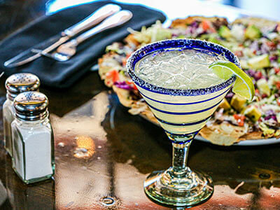 Copa de margarita con rodaja de lima sobre mesa, cerca de un plato de nachos y salero y pimentero.