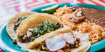 Tres tacos con carne, cilantro y rábanos, acompañados de arroz, frijoles y plátano en un plato azul.