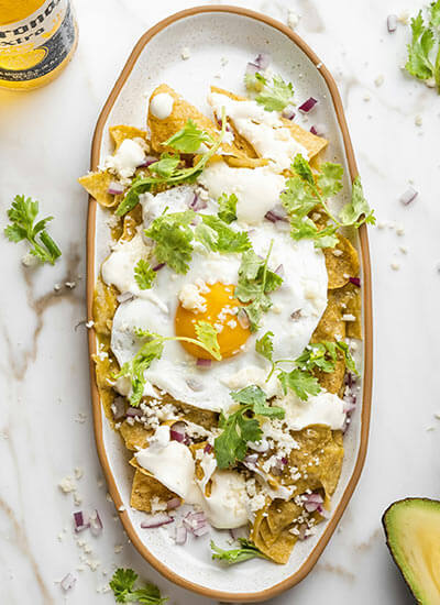 Plato ovalado de chilaquiles verdes con huevo estrellado, crema, queso, cebolla morada y cilantro. Botella de cerveza al lado.