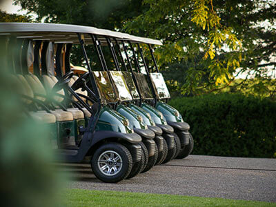 Varios carritos de golf alineados en un camino, rodeados de árboles y césped, bajo luz natural.