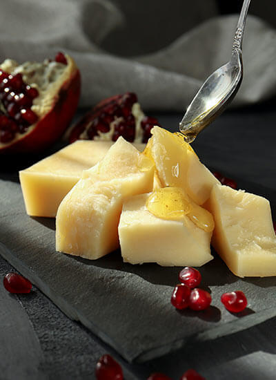 Trozos de queso con miel siendo vertida, rodeados de granos de granada sobre una superficie de pizarra.