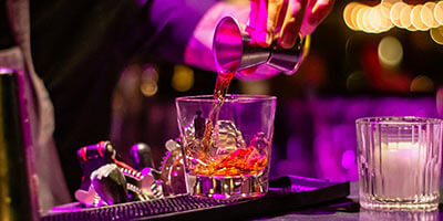 Persona sirviendo cóctel de licor en un vaso con fondo iluminado en tonos púrpura.