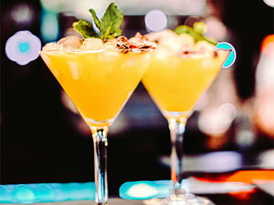 Dos cócteles amarillos con hielo y hojas de menta en copas de martini sobre una barra, fondo desenfocado.