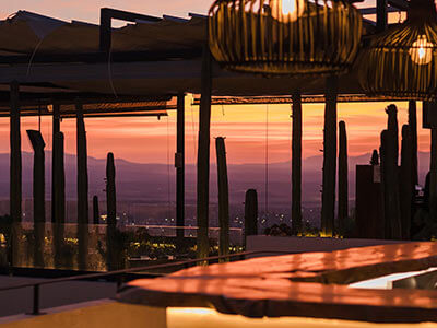 Terraza al atardecer con lámparas colgantes y vista a un paisaje montañoso con cactos silueteados.