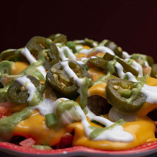 Nachos con queso derretido, jalapeños, crema agria y guacamole en un plato rojo sobre fondo oscuro.